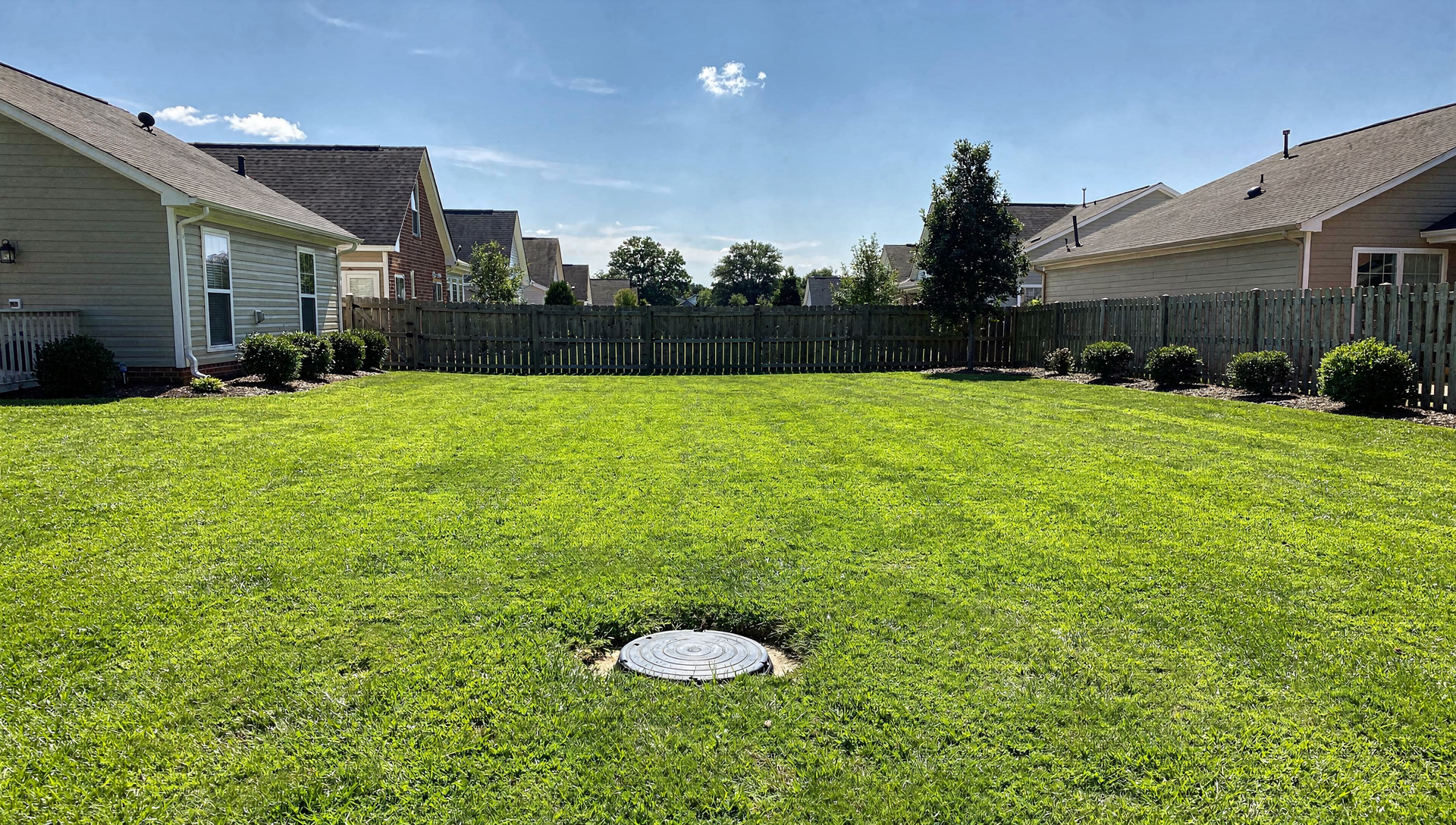 A bright Charlotte back yard with a healthy green lawn and a septic access lid in the grass under a blue sky